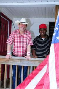 Two men smiling at the porch of their home. One has a cowboy hat and plaid shirt, the other a red baseball cap and striped polo.