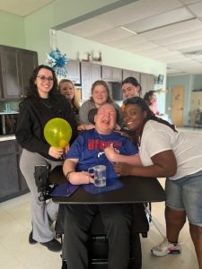 Liberty ARC Direct Support Professionals stand around smiling man in wheelchair with balloon for birthday.