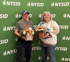 Donna Vincent, the Horticulture Coordinator, and Roger Foster, an employee of the Horticulture Center in front of a NYSID backdrop holding floral arrangements.